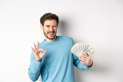 Cheeky smiling man winking, showing Ok sign and holding money, concept of fast loan or credit, standing over white background.