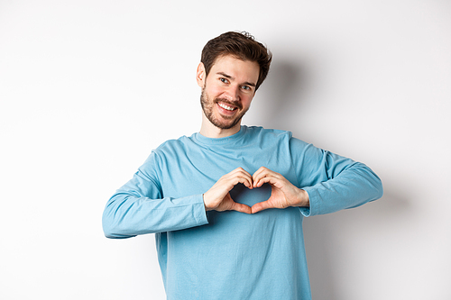 Handsome boyfriend saying I love you, showing heart gesture and smiling at camera, express love and romantic feeling, standing over white background.