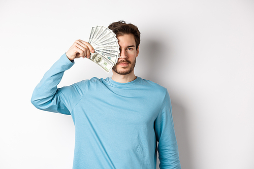 Serious young man covering half of face with money, looking at camera, standing in blue sweatshirt over white background.
