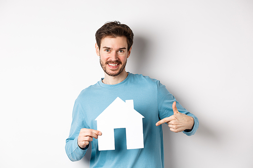 Real estate and insurance concept. Smiling young man pointing at paper house cutout and looking happy, buy apartment, standing over white background.