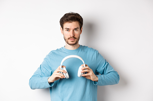 Young bearded guy in blue sweatshirt put on wireless headphones, listening music, standing on white background.