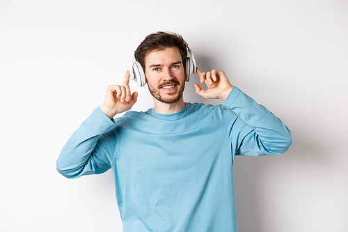 Attractive modern man with beard, smiling pleased, touching headphone on head while listening to music, standing over white background.