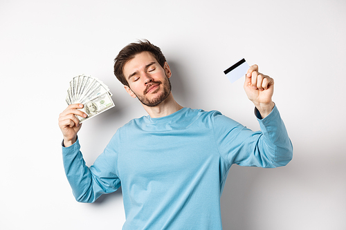 Carefree male model dancing with money and credit card, smiling and dreaming of shopping, standing over white background.