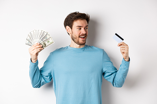 Handsome bearded man choosing between money and plastic credit card, payment with cash or contactless, standing over white background.