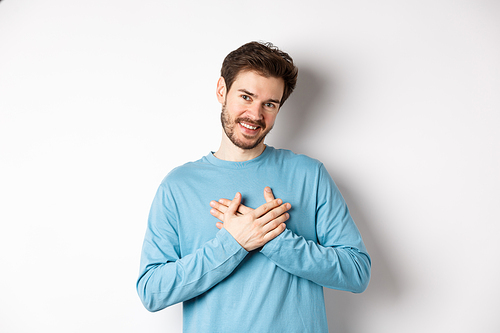 Portrait of young handsome man with beard, holding hands on heart and saying thank you, thinking about pleasant moment, standing over white background.