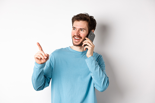 Technology concept. Young man talking on mobile phone and pointing left, having call on smartphone, standing over white background.