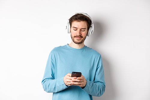 Modern young man in blue sweatshirt put on music on smartphone, listening songs in wireless headphones, white background.