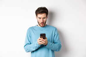 Amazed young man staring at smartphone screen and saying wow, looking at online offer, standing on white background.