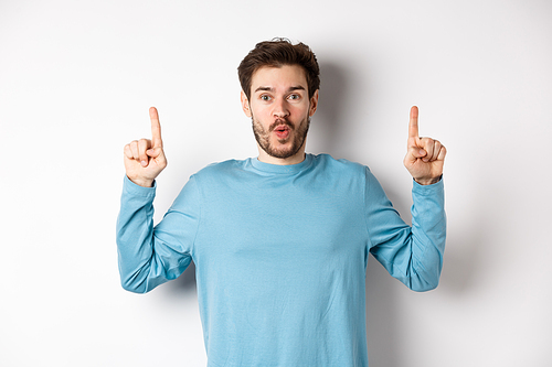 Surprised young man showing top advertisement, pointing up and saying wow at camera, standing over white background.