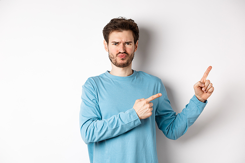 Disappointed and upset young man showing bad offer, pointing fingers right and frowning disgusted, standing with skeptical face on white background.