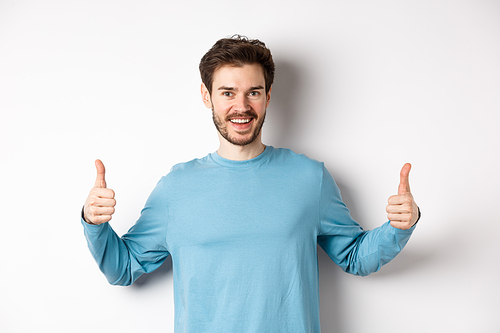 Cheerful young man showing thumbs up and smiling, recommend good product, praising choice, standing on white background.