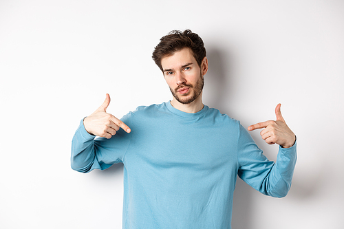 Young man with beard feeling confident, pointing at himself and making smug face, standing on white background cool and sassy.