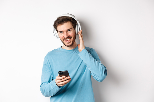 Good-looking guy listening music in wireless headphones, smiling at camera and using smartphone, standing on white background.