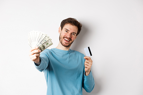 Handsome young man in blue shirt giving you money, holding plastic credit card, standing over white background.