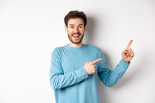 Cheerful smiling man pointing fingers right and showing banner or logo on white space for advertisements, recommending product, standing over studio background.