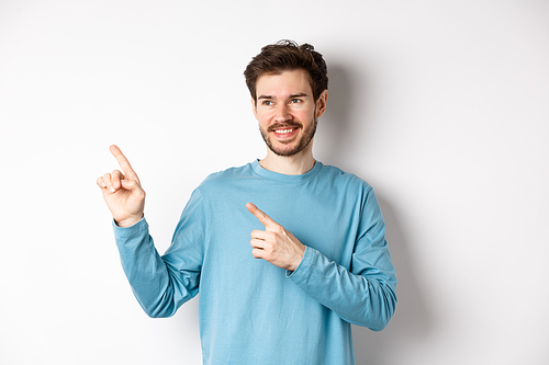 Portrait of young modern man in blue sweatshirt pointing fingers at upper left corner and looking away, smiling pleased as showing advertisement on white background.