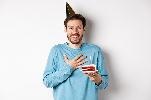 Celebration and holidays concept. Surprised birthday boy in party hat, holding bday cake and looking thankful, standing over white background.