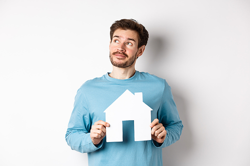 Real estate and insurance concept. Man dreaming about buying property, holding paper house cutout and looking at upper left corner, imaging things, white background.