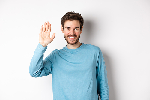 Cheerful young man with beard saying hello, looking friendly and waving hand to greet you, standing over white background.