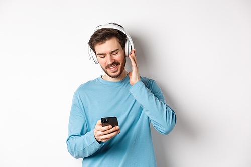 Cheerful smiling man listening music and looking at smartphone, reading message on phone, standing on white background.