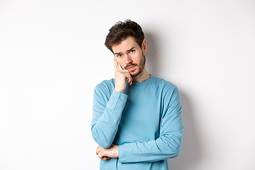 Skeptical young man listening to you with bored face, looking at camera reluctant, standing in sweatshirt over white background.