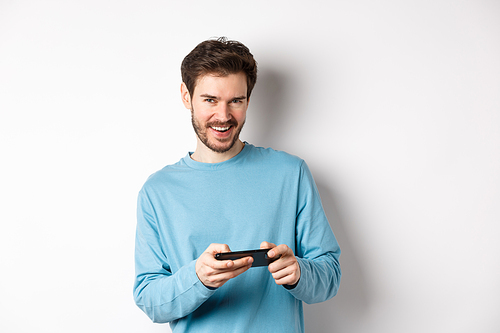 Image of handsome young man playing mobile video game, holding smartphone horizontally and smiling at camera pleased, standing over white background.