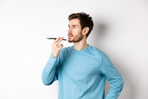 Handsome young man recording voice message on smartphone, holding phone near lips and talking. Guy using translator app on mobile phone, white background.