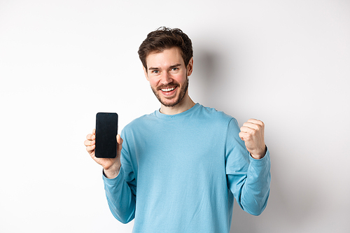 Image of happy man showing empty smartphone screen and celebrating, smiling with rejoice and fist pump, achieve online goal, standing over white background.