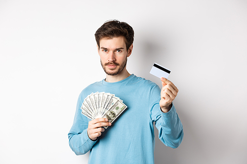 Caucasian man holding money and showing plastic credit card, standing in blue sweatshirt on white background.