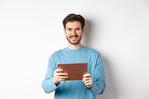 Handsome young man holding digital tablet and smiling, networking with device, standing over white background. Copy space