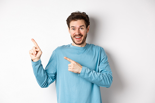 Happy caucasian man in casual blue sweatshirt, pointing fingers at upper left corner, showing link or logo on white background, smiling at camera.