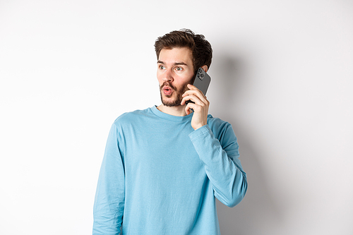 Technology concept. Intrigued young man looking left impressed, talking on mobile phone, calling someone, standing over white background.