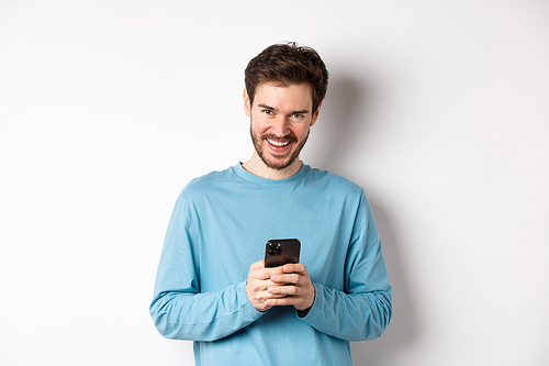 Image of handsome man using smartphone and laughing, smiling at camera joyful, standing over white background.