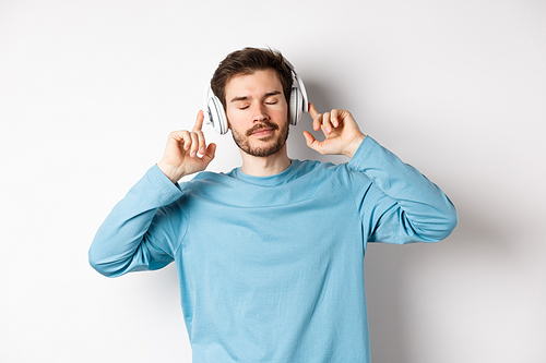 Relaxed young man enjoying favorite song, listening music in headphones with closed eyes and calm face, standing over white background.