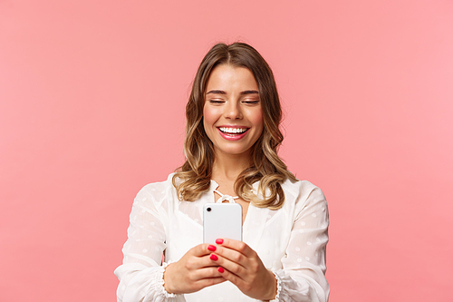 Close-up portrait of happy charming caucasian woman with short fair hair, white dress, record video of concert on mobile phone, taking photo using smartphone, standing pink background.