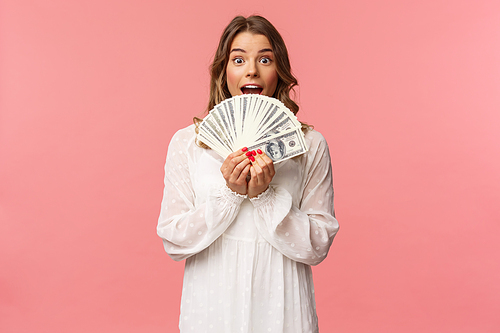 Portrait of excited lucky girl with blond hair, white dress, winning money, receive cash award, big lottery prize, holding dollars near face, smiling and looking amazed, standing pink background.