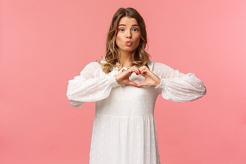 Beauty, fashion and women concept. Portrait of tender lovely young blond girl in beautiful white dress, fold lips in kiss and make heart sign near chest, standing pink background, show sympathy.