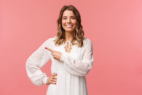 Pleasant good-looking smiling blond woman with blond short wavy hair, white tender dress, looking camera and pointing finger left to show you best price, good offer, promo concept, pink background.