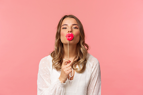 Spring, happiness and celebration concept. Close-up of romantic attractive blond caucasian girl in white dress, holding big kissing lips stick over mouth and looking lovely camera, pink background.