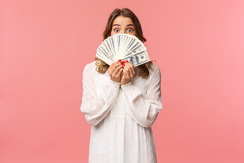 Portrait of amazed and excited cute feminine blond girl in white dress, holding dollars over face, looking from under cash at camera with surprised expression, stand pink background.