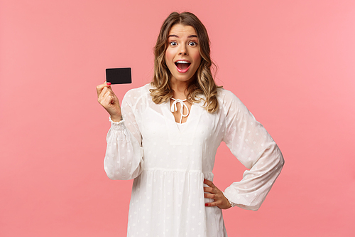 Portrait of happy excited good-looking blond girl in white dress, being impressed and pleased with cool banking service, showing credit card, smiling camera, standing pink background.