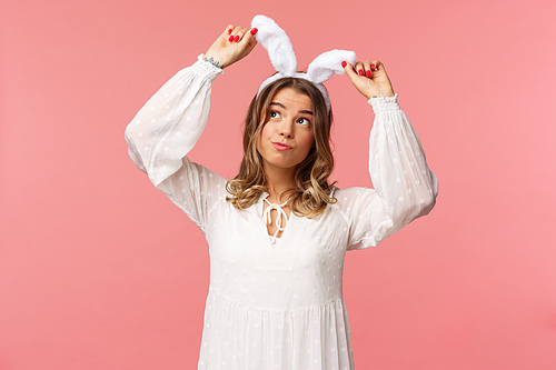 Holidays, spring and party concept. Portrait of silly cute blond girl in rabbit ears, looking up daydreaming, smiling lovely wear white dress, standing over pink background carefree.