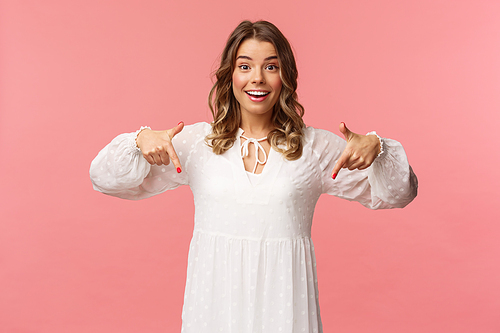 Portrait of enthusiastic upbeat young blond woman pointing fingers down to invite you check-out product, showing bottom advertisement, smiling camera cheerful, spring concept, pink background.