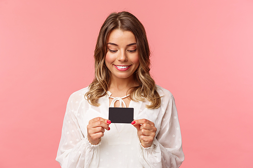 Close-up portrait of excited and amused blond girl in white dress, holding credit card and smiling thrilled, cant resist temptation to buy something, waste money online shopping, pink background.