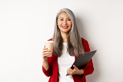 Successful asian businesswoman drinking coffee and smiling, standing with clipboard, wearing red blazer, white background.