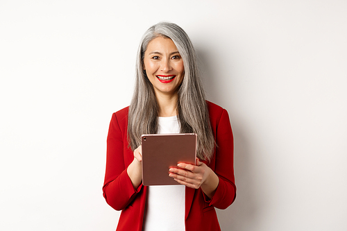 Business. Senior female entrepreneur working on digital tablet and smiling happy at camera, wearing red blazer and makeup, standing over white background.