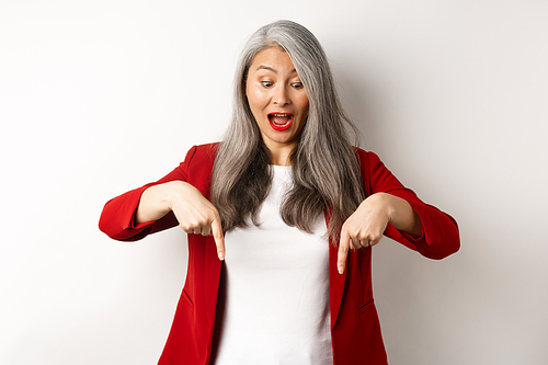 Stylish asian elderly woman in red blazer checking out special promotion, pointing fingers down and looking excited, scream of joy, white background.