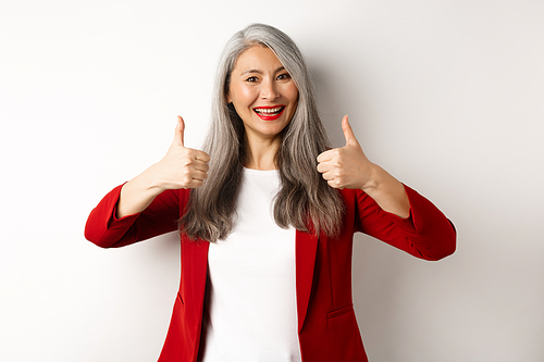 Portrait of senior asian woman showing thumb-up in approval, wearing red blazer for office work, recommend company, standing over white background.