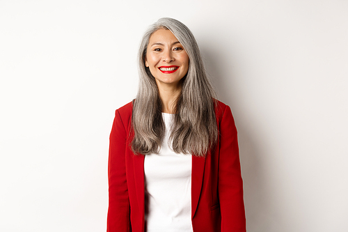 Business people. Senior smiling asian woman with red lipstick and blazer looking happy, standing over white background.