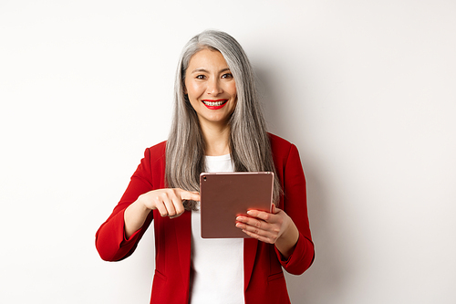 Business. Smiling senior businesswoman using digital tablet, pointing finger at screen, standing over white background.